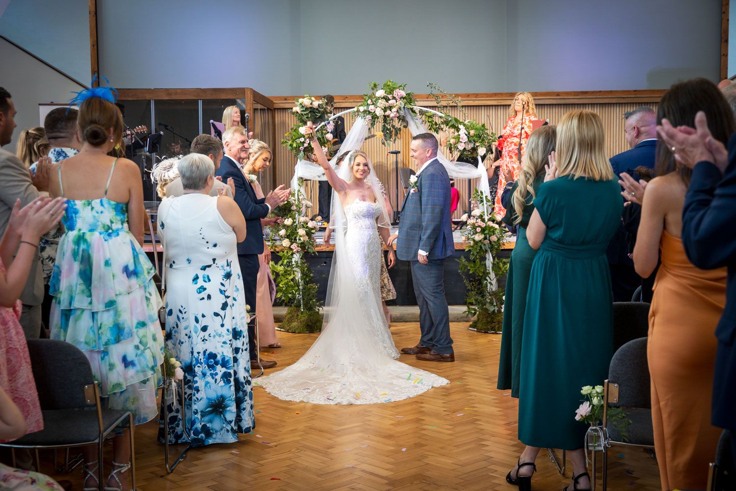 Wedding guests and attendees watch a ceremony in a beautifully decorated reception hall with floral arrangements.