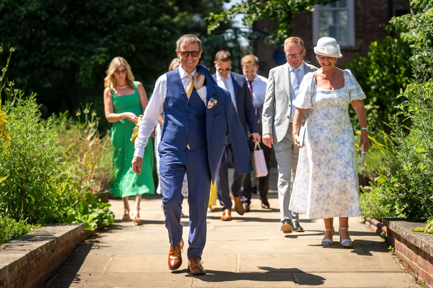Wedding guests walking along a sunny garden path in smart formal attire.