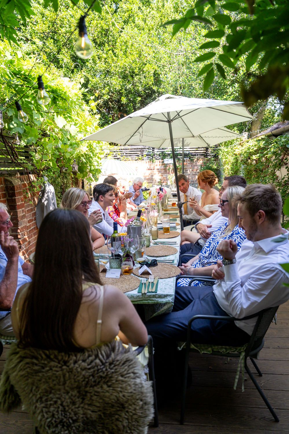 Wedding reception guests dining at a long table in a lush garden setting with festoon lights.