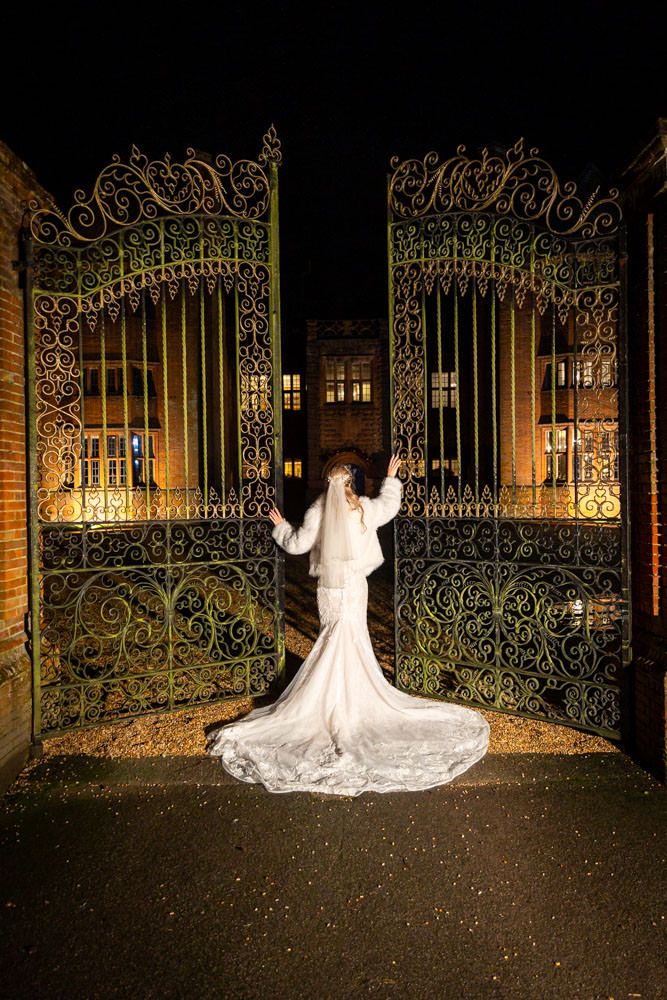 Bride in white gown with train standing before ornate iron gates at night, New Place hotel behind