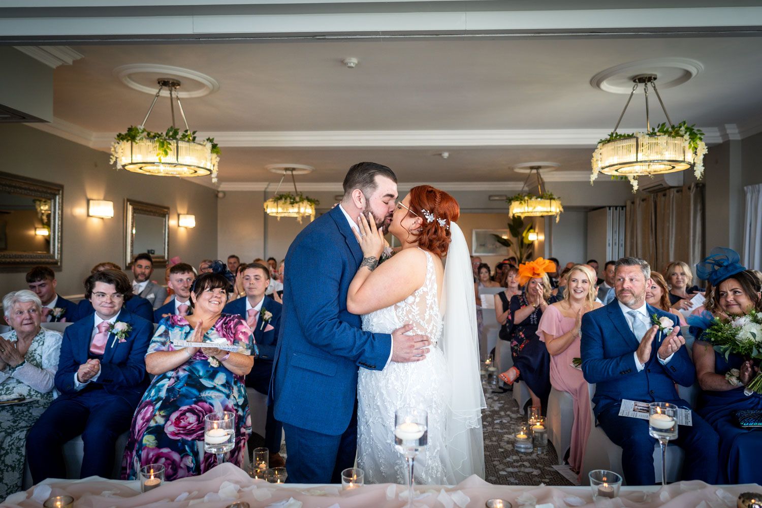 Newlywed couple shares their first kiss at wedding reception while guests applaud in elegant ballroom with chandeliers.