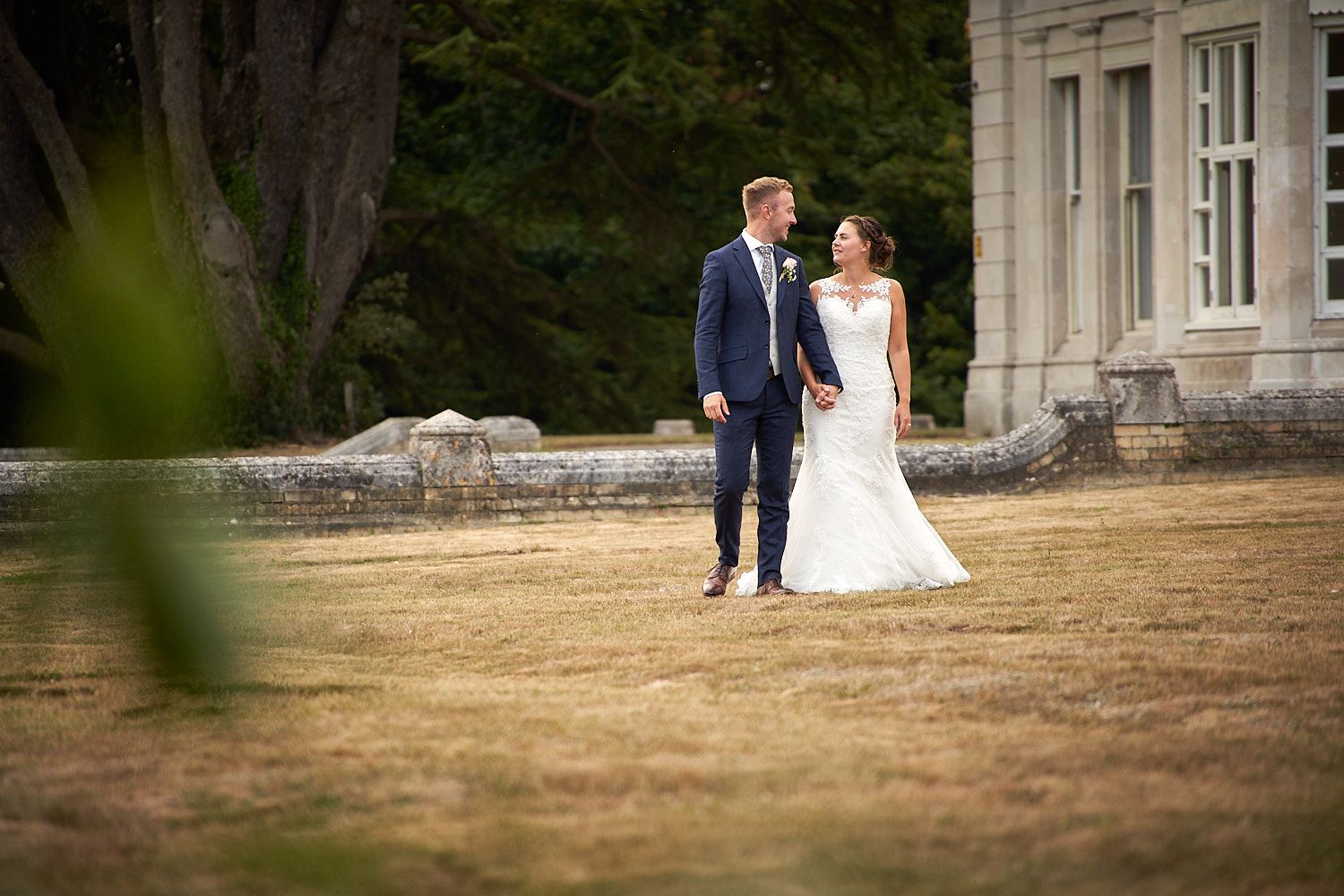 Happy couple poses in wedding attire on a dry lawn in front of a stately white building during summer.