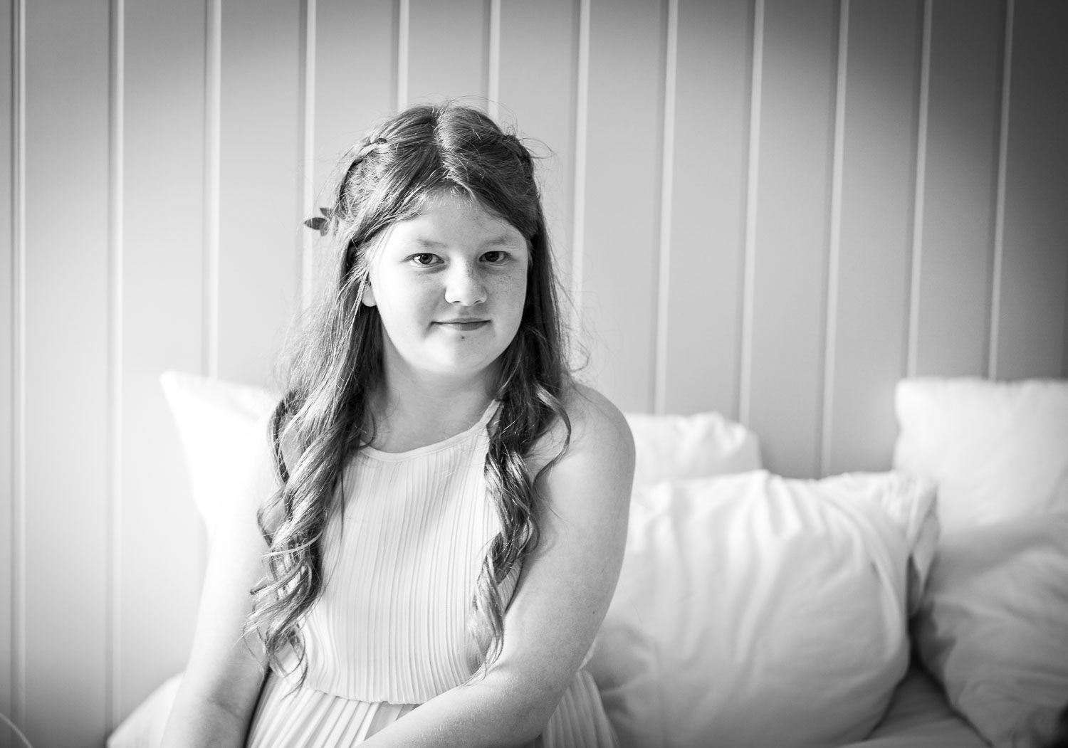 Black and white portrait of a subject with long wavy hair wearing white clothing while sitting on a bed.