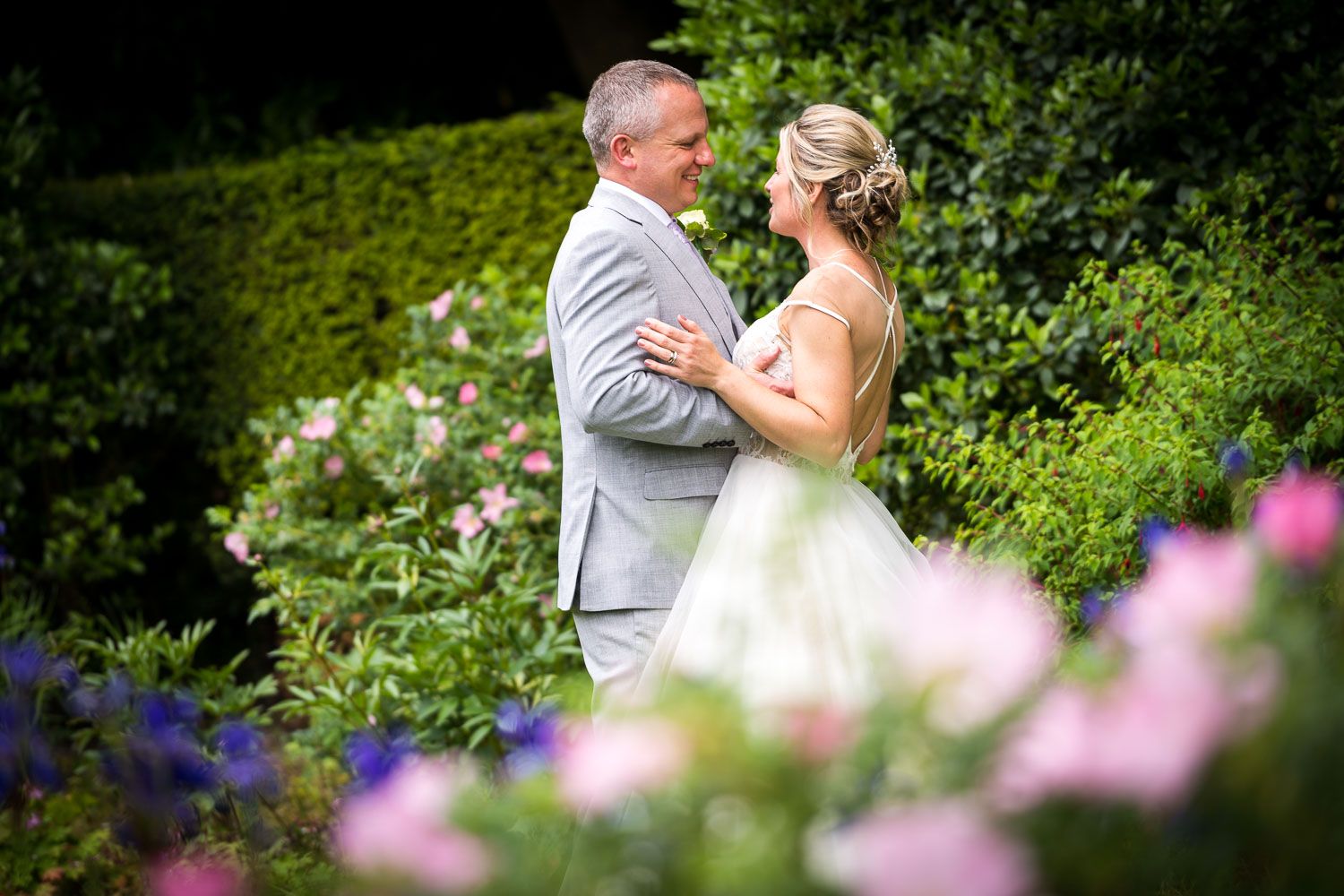 A bride and groom embrace in a garden setting surrounded by colorful summer flowers and lush greenery.