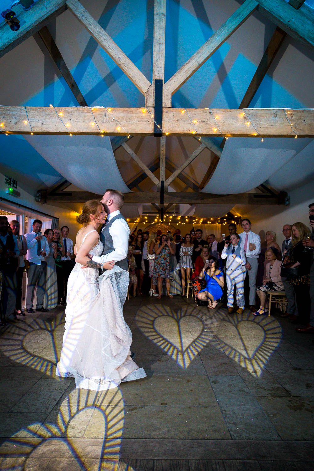 A couple shares their first dance at a wedding reception under a beam ceiling decorated with string lights and pattern lighting.
