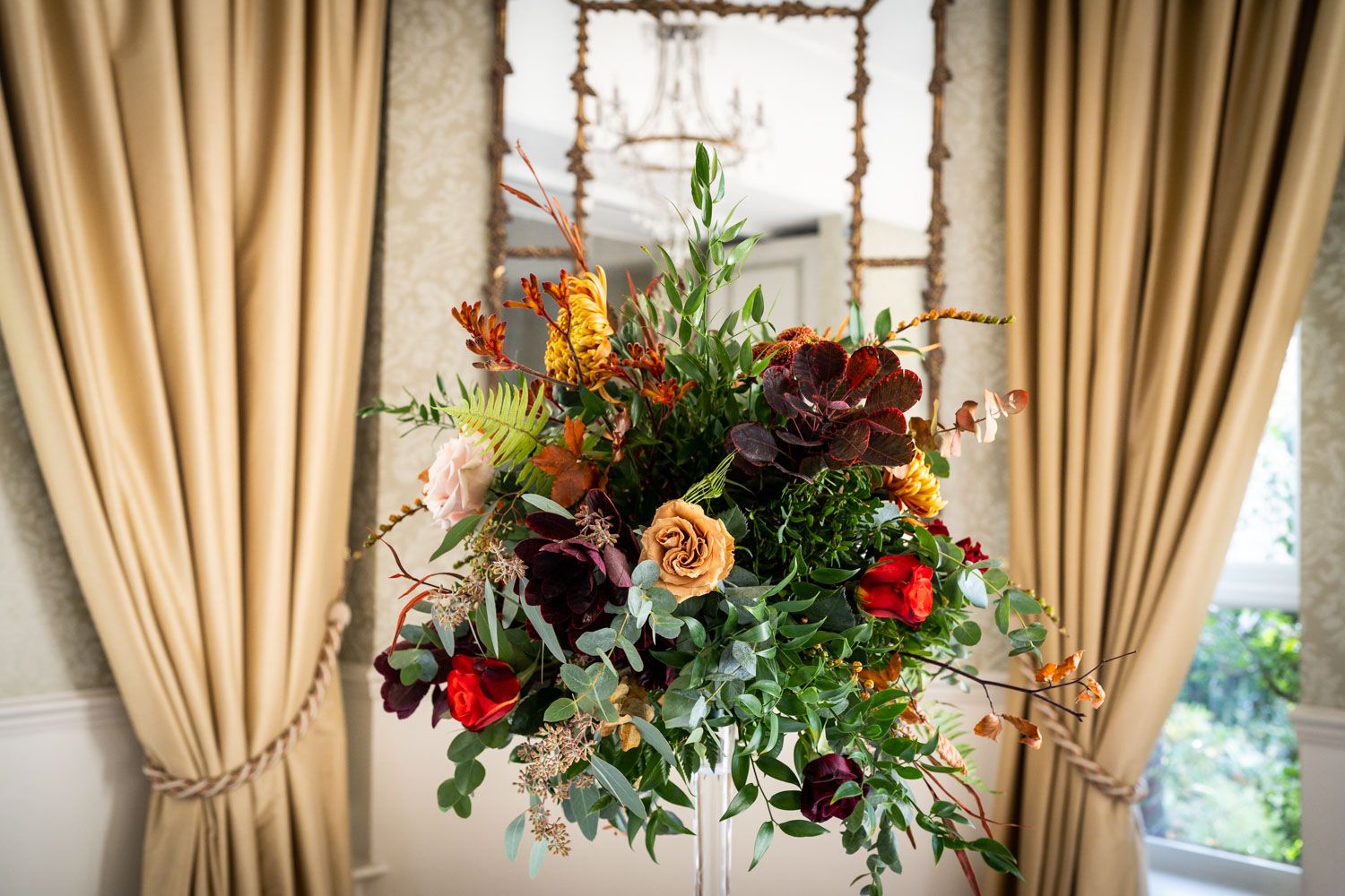 A stunning autumn floral arrangement with roses, berries, and greenery hangs elegantly against beige curtains by a window.