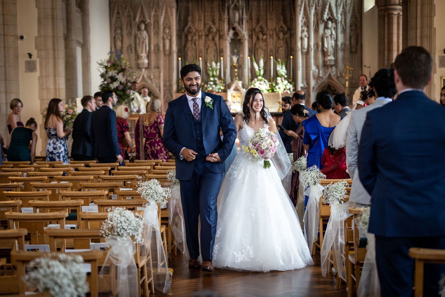 Elegant wedding ceremony takes place inside a grand gothic cathedral with wooden pews decorated with floral arrangements.