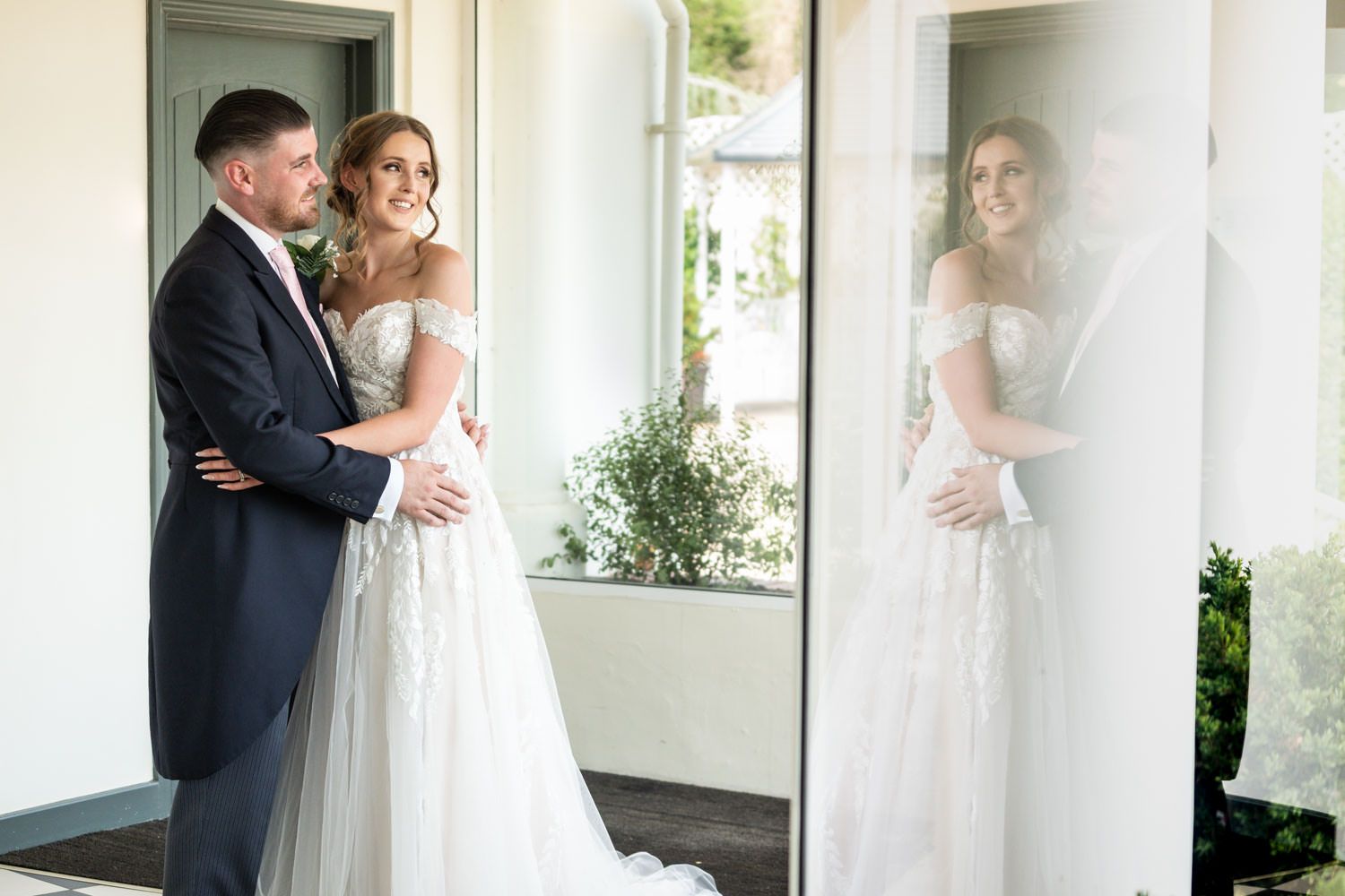 A couple stands together in an elegant wedding photo with a reflection in the window glass.