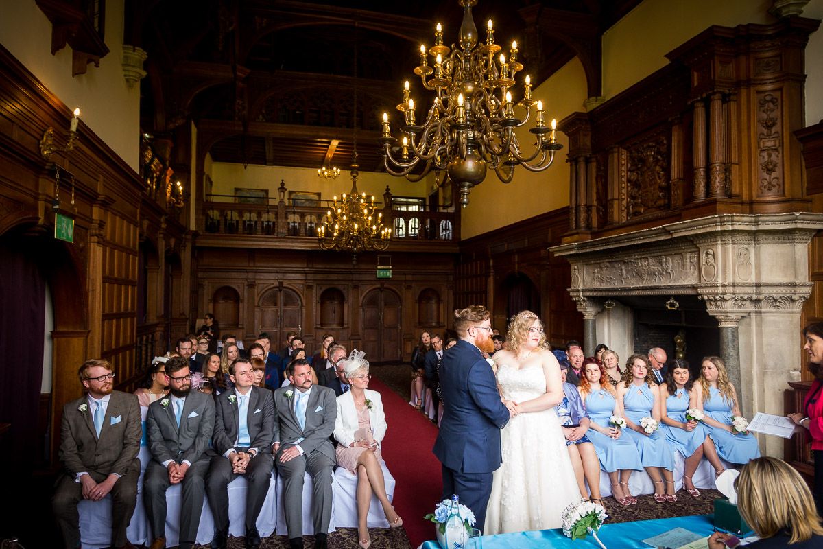 Wedding ceremony in elegant historic hall with ornate chandelier, stone fireplace and wedding party in blue and grey attire.