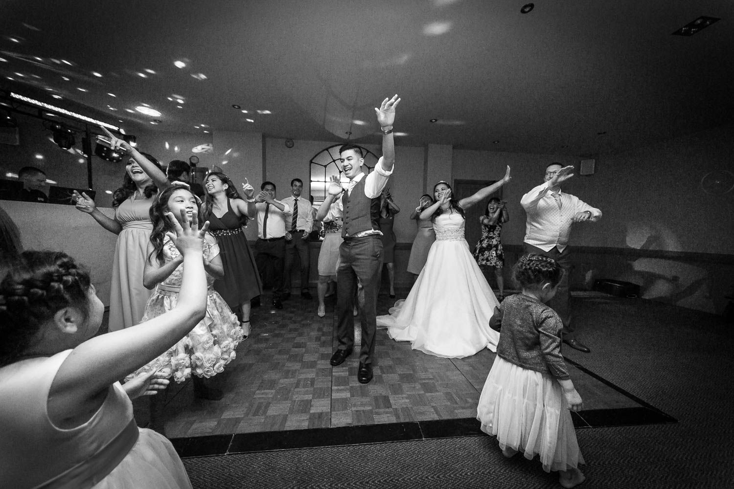 Wedding party guests celebrate and dance energetically on a reception dance floor in black and white photography.