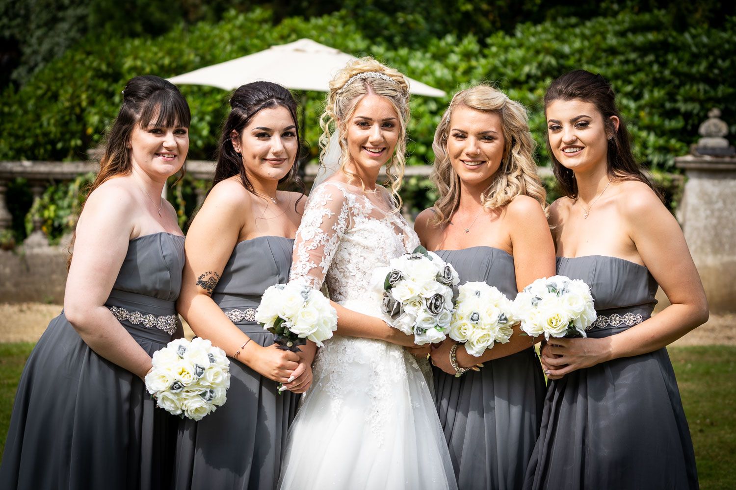 Wedding party poses outdoors holding white bouquets with bridesmaids in gray dresses and bride in white gown.