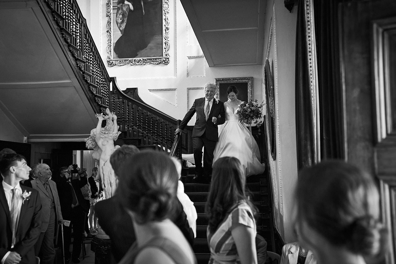 Wedding guests gather at an elegant staircase during a formal celebration captured in black and white photography.