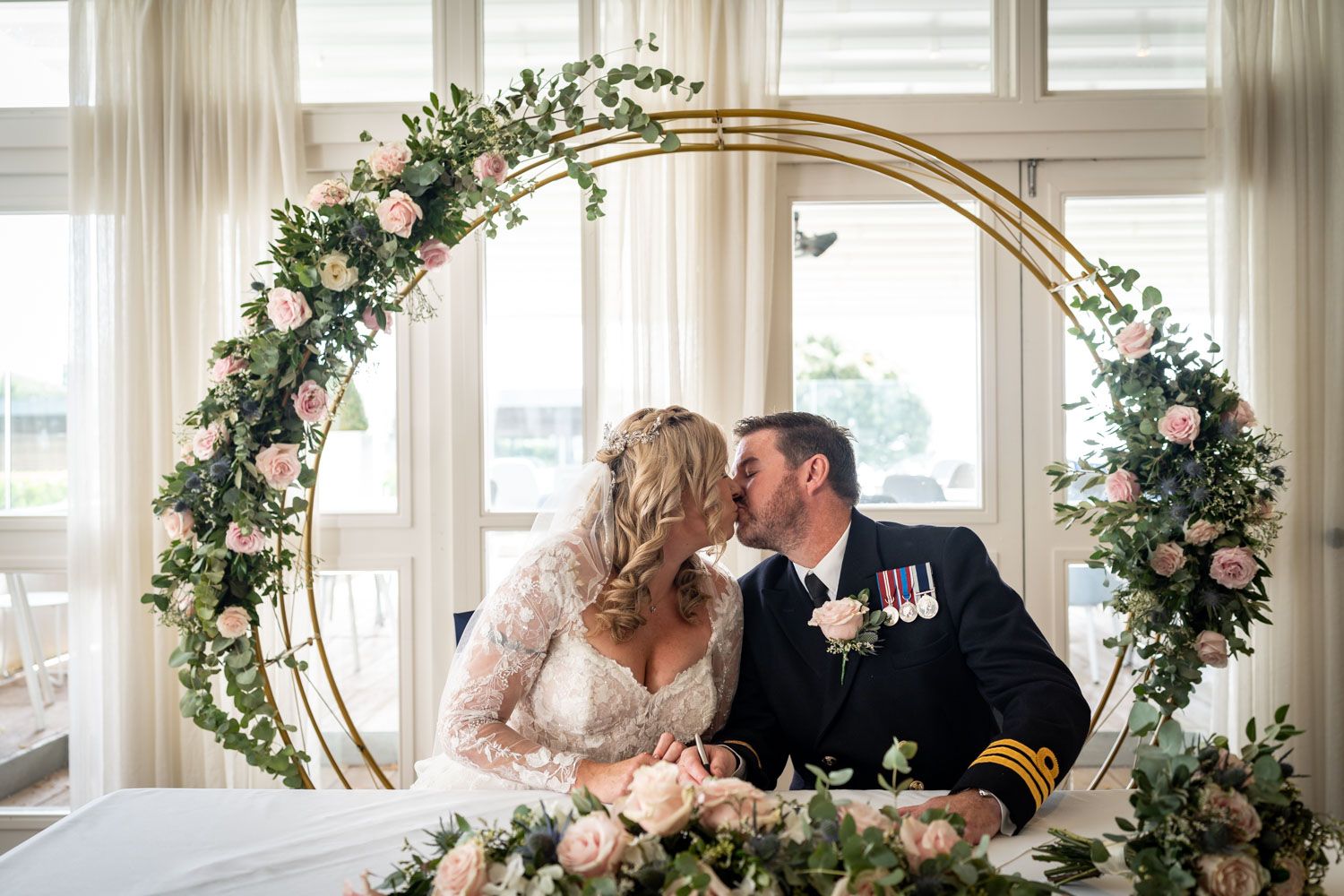 Romantic wedding photo with a couple sitting under a circular floral arch decorated with pink roses and greenery.