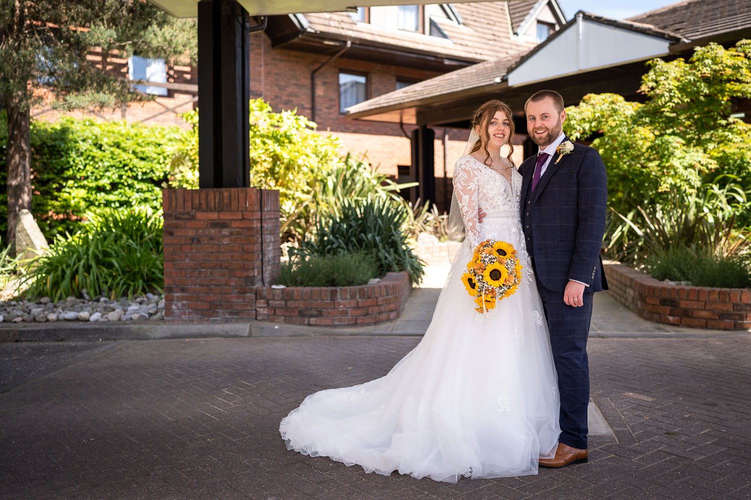 A couple poses at their wedding ceremony with yellow sunflower bouquet in front of a brick garden entrance.