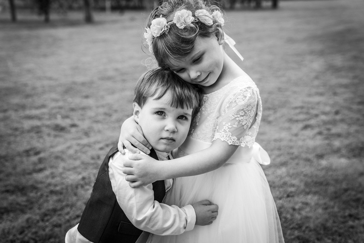 A young boy in a suit and a girl in a white dress with a flower crown share a tender embrace in black and white.