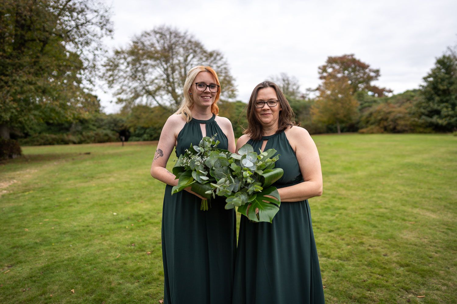 Two people in matching dark green bridesmaid dresses hold eucalyptus bouquets while standing on a lawn.