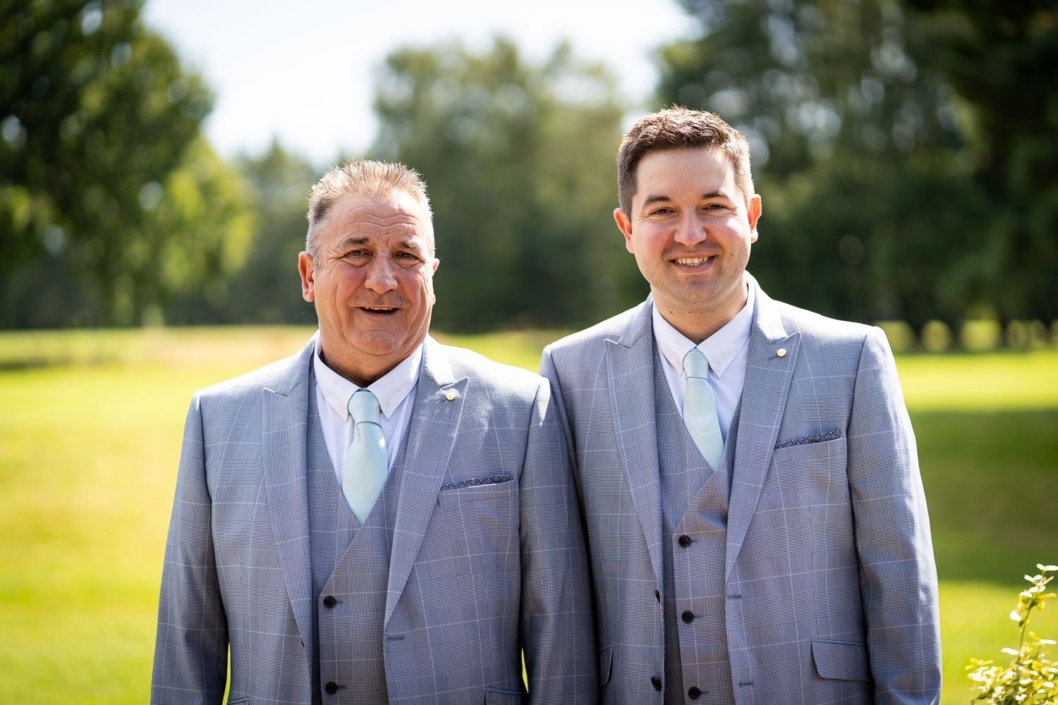 Two people wearing matching gray suits and light blue ties stand together smiling outdoors on a sunny day.