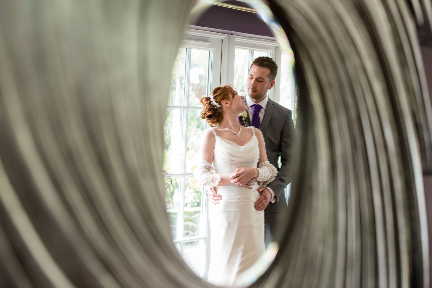 Wedding couple seen through mirror reflection in elegant indoor setting with natural window light.