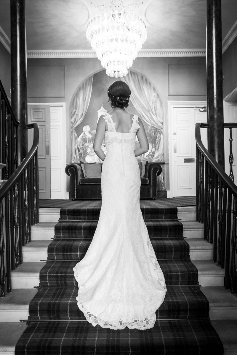 A stunning black and white photo of a bride in a fitted lace wedding gown standing on a grand staircase under a chandelier.