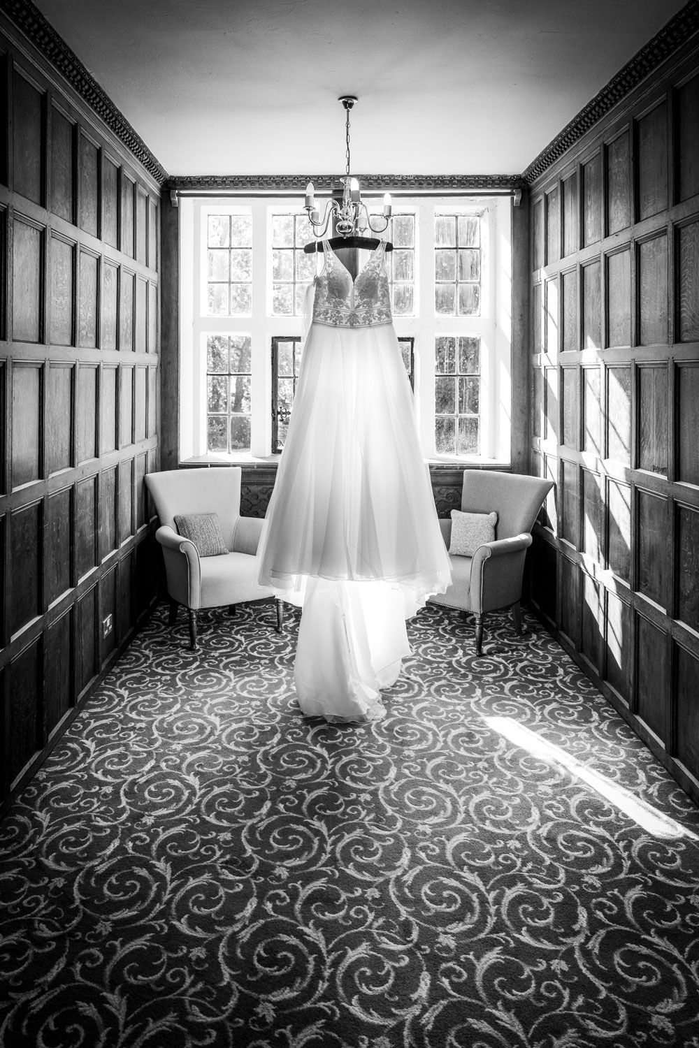 Elegant wedding dress hanging from a chandelier in a wood-paneled room in black and white.
