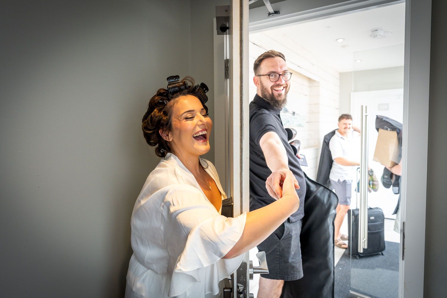 Bride laughing joyfully in white robe as groom arrives at hotel room door before wedding.