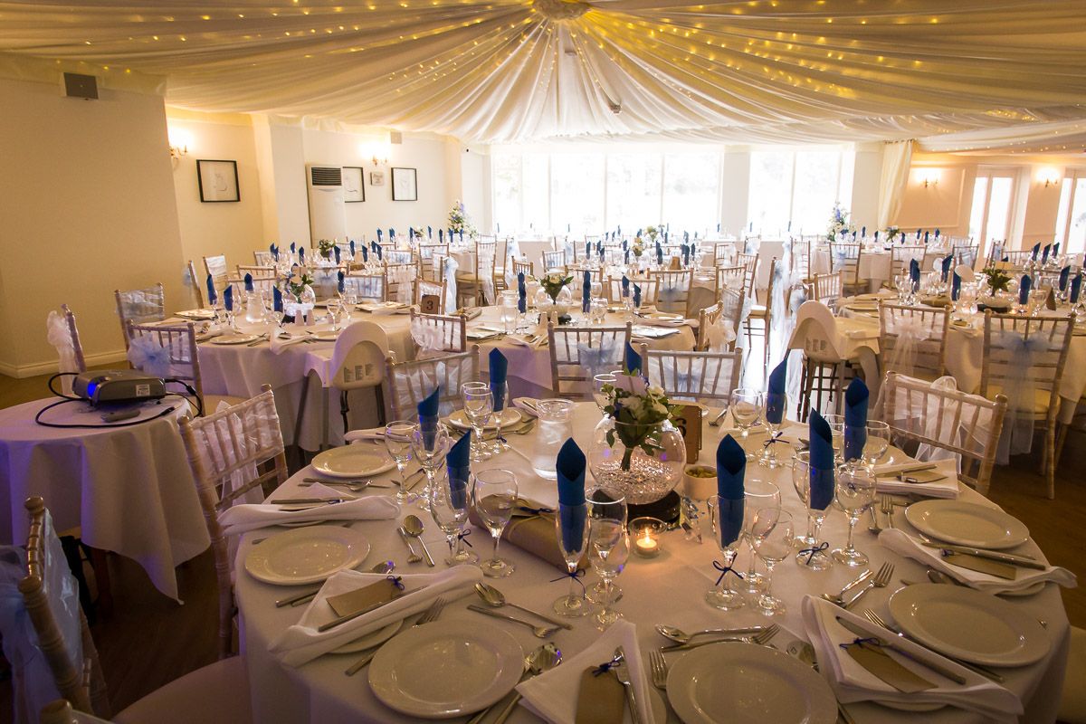 An elegant wedding reception venue with round tables, white linens, draped ceiling, and purple accents throughout the room.