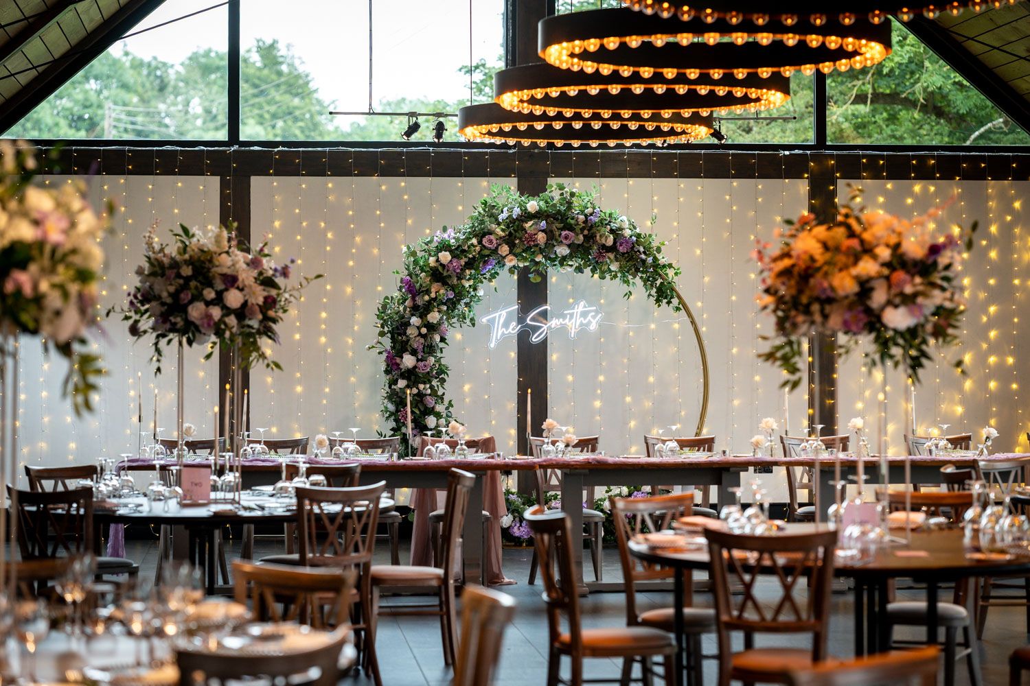 Elegant wedding reception venue with floral arch, string lights, and dark wood tables in a modern glass-enclosed space.