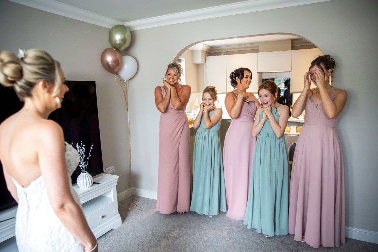 Bridesmaids in pink and mint dresses react emotionally to seeing bride in wedding gown at bridal suite before ceremony.