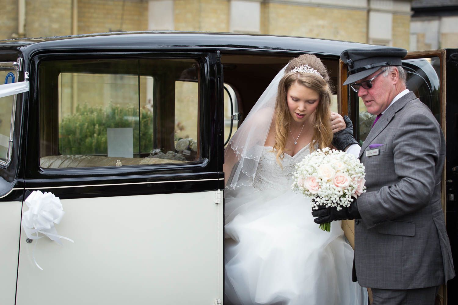 A bride in a white dress holding a bouquet steps into a classic vintage wedding car assisted by someone in gray.
