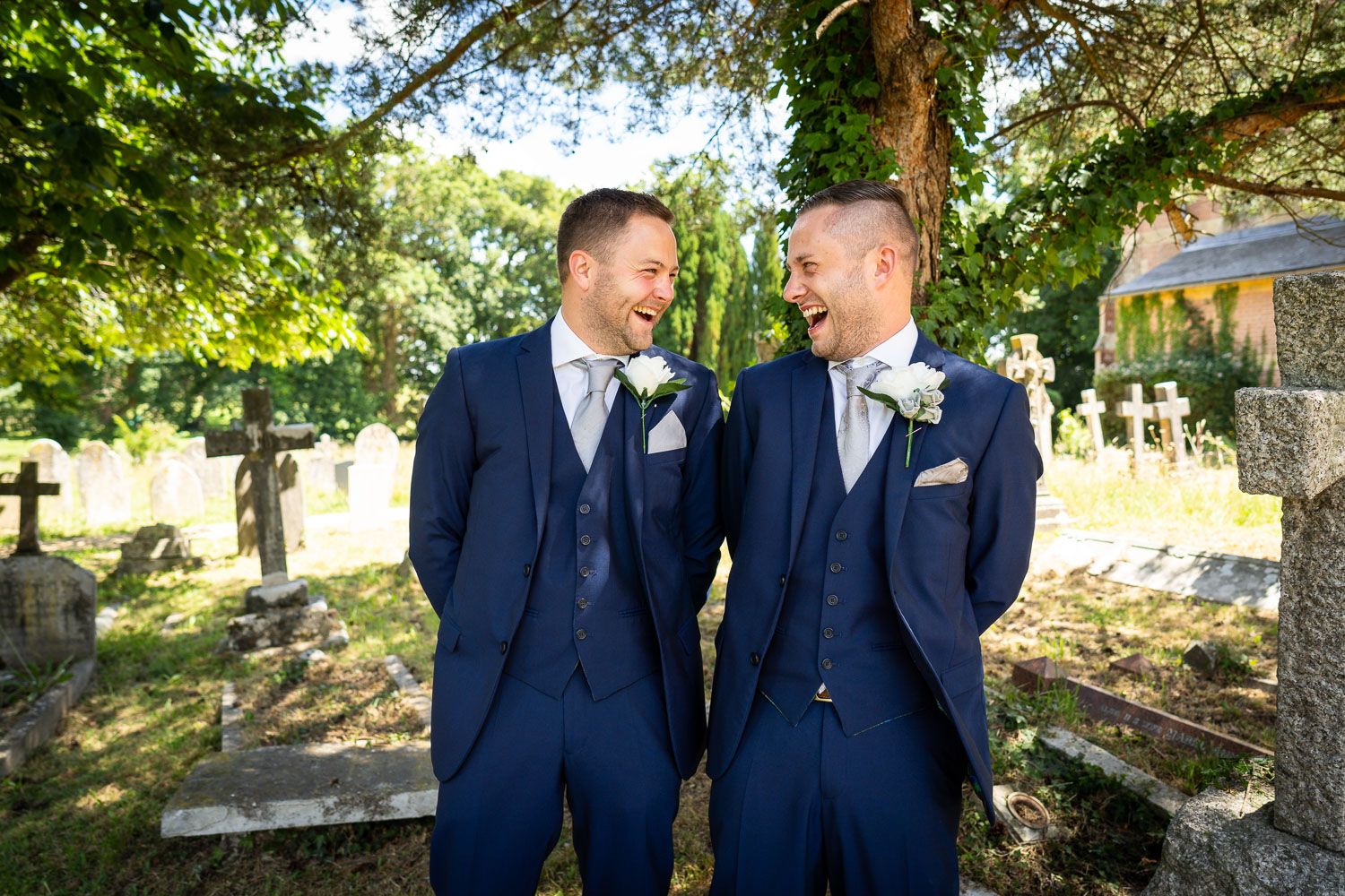 Two people in matching navy blue three-piece suits smile at each other outdoors under trees at a wedding ceremony.