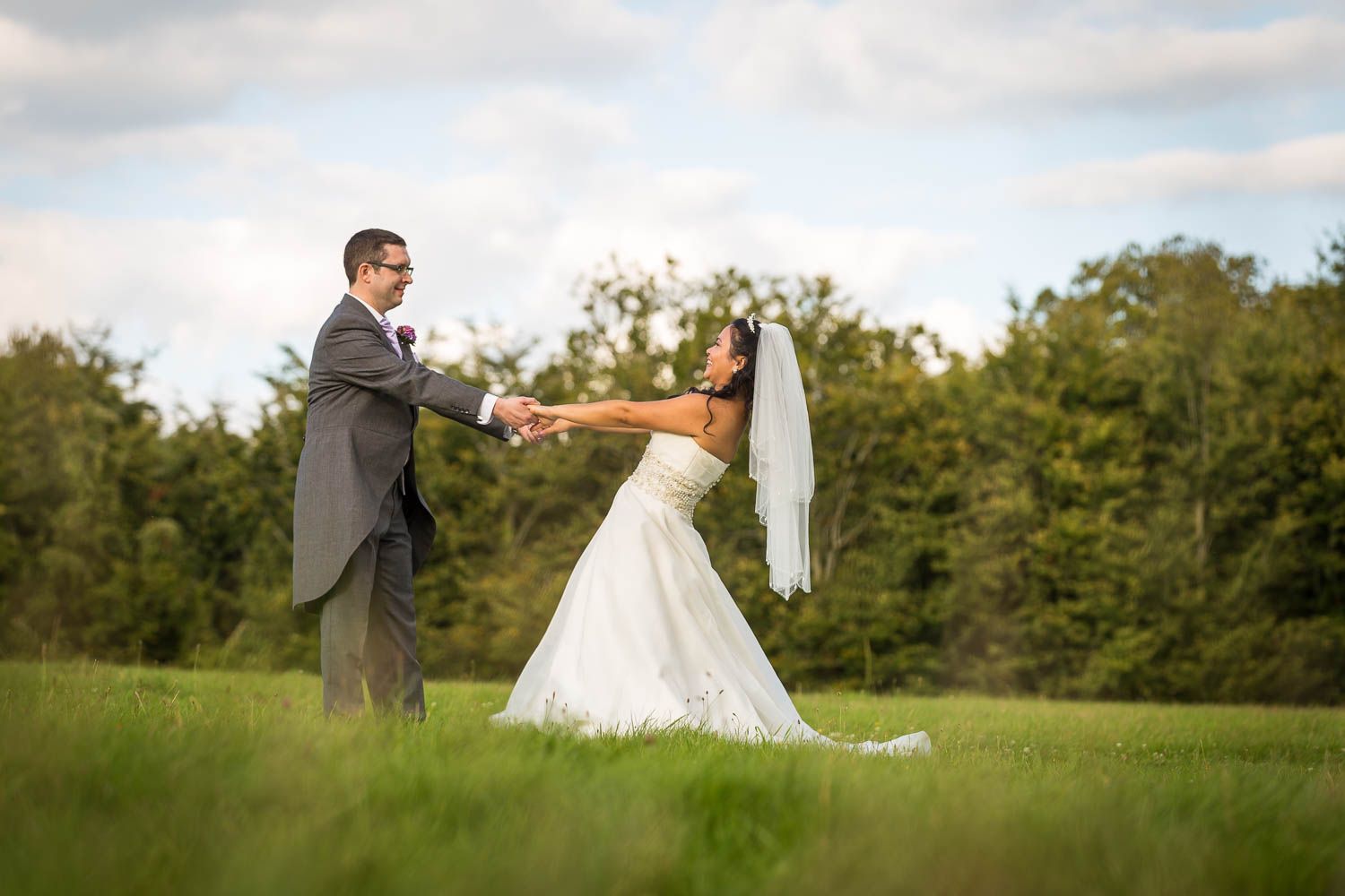 A couple dances and twirls together on a grassy field during their wedding celebration with trees in the background.