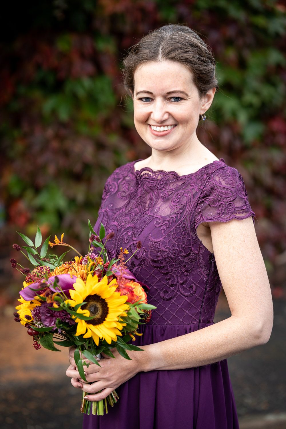 Someone in a purple lace bridesmaid dress holds a vibrant bouquet with sunflowers and fall flowers against autumn foliage.