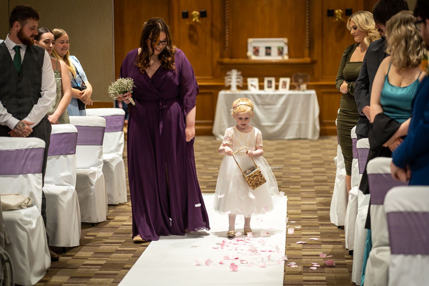 A small child in a white dress walks down a rose petal-covered aisle while guests watch during a wedding ceremony.