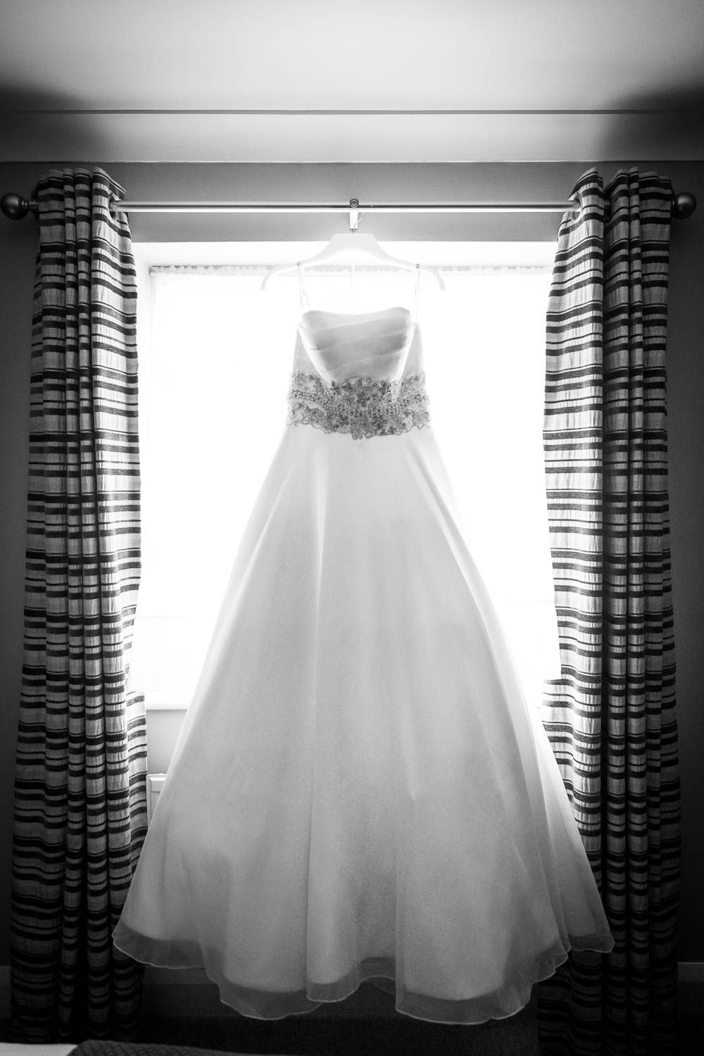 A white wedding dress with beaded bodice hangs elegantly in front of a window framed by striped curtains in black and white.