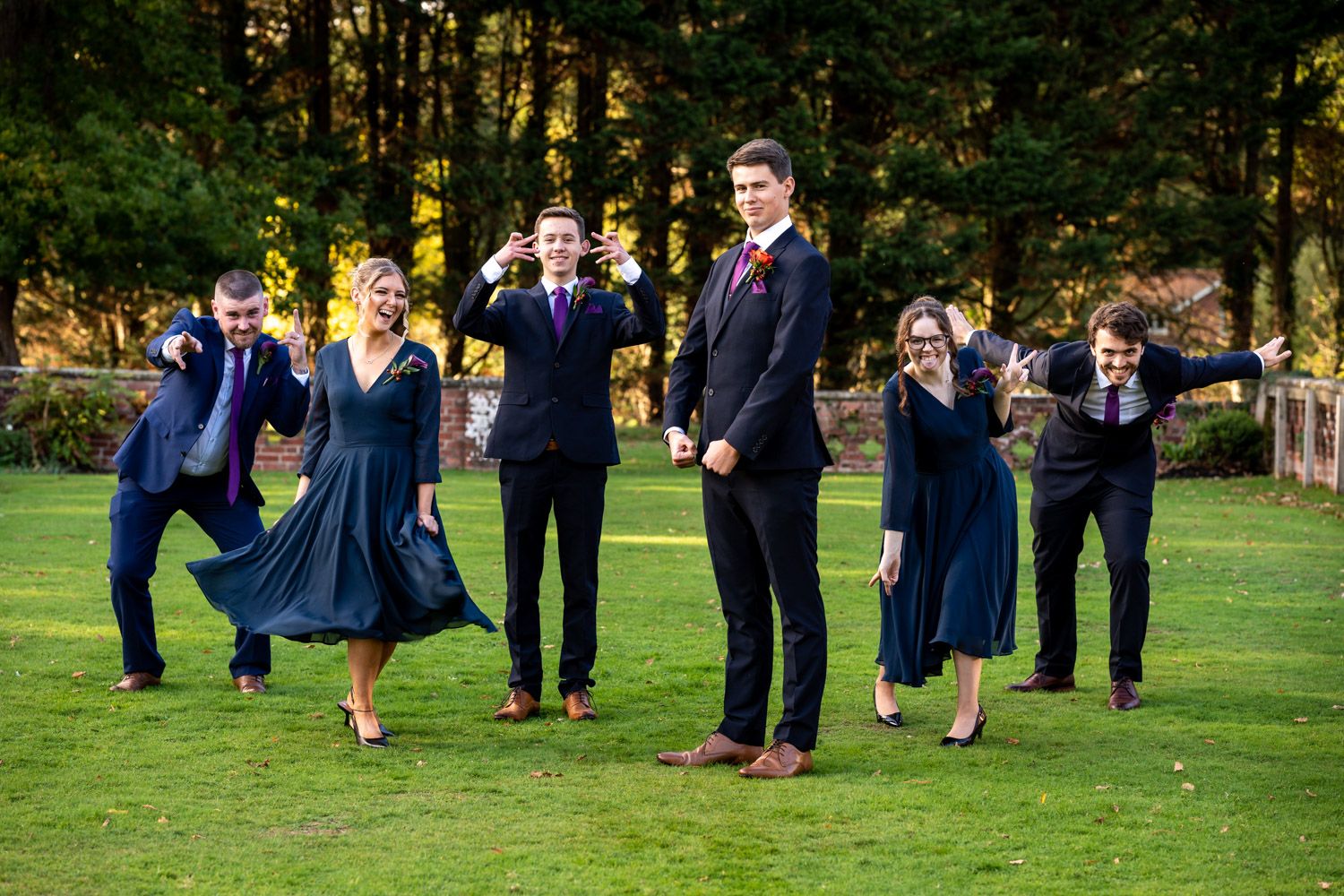 A wedding party in navy blue and black attire poses playfully on a green lawn with forest backdrop.
