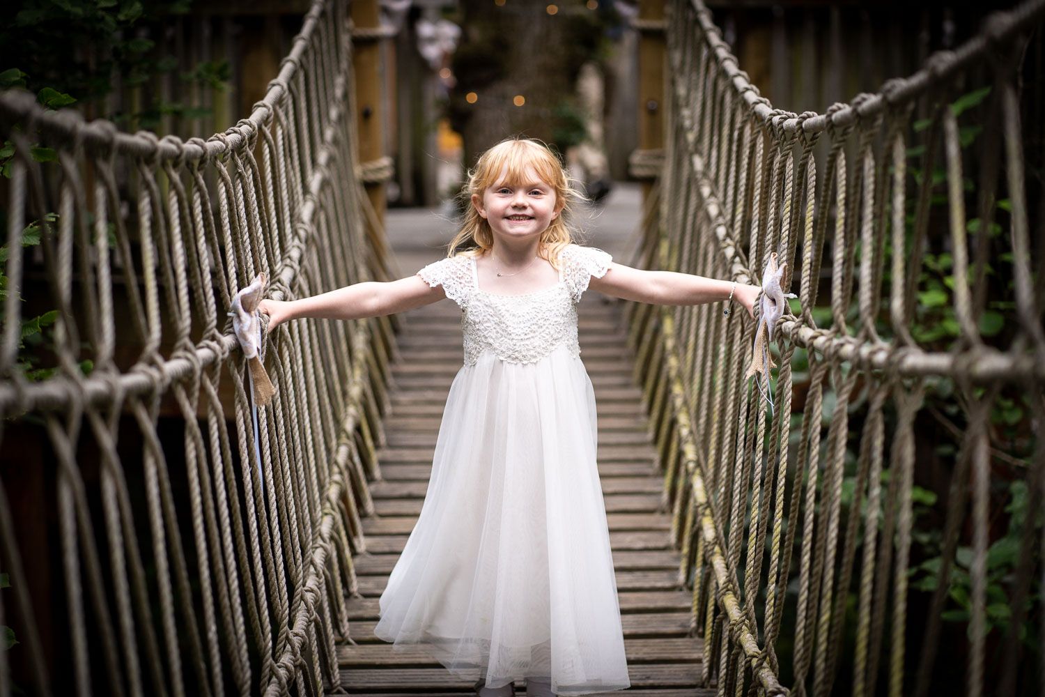 A young child in a white dress spreads their arms while standing on a rope bridge in nature.