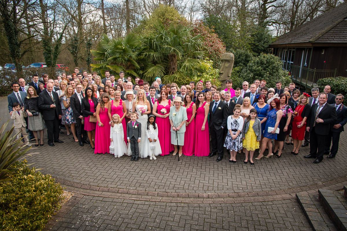 A large wedding party poses together on a brick patio surrounded by garden landscaping in formal attire.