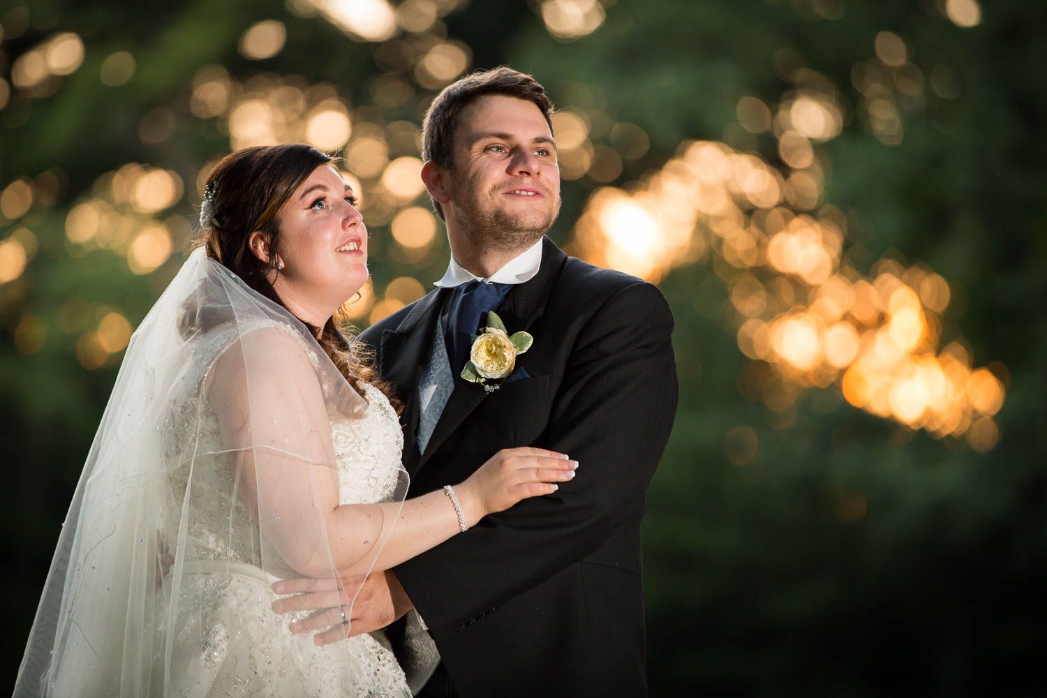 A bride and groom share a romantic moment at sunset with bokeh lights glowing in the background.