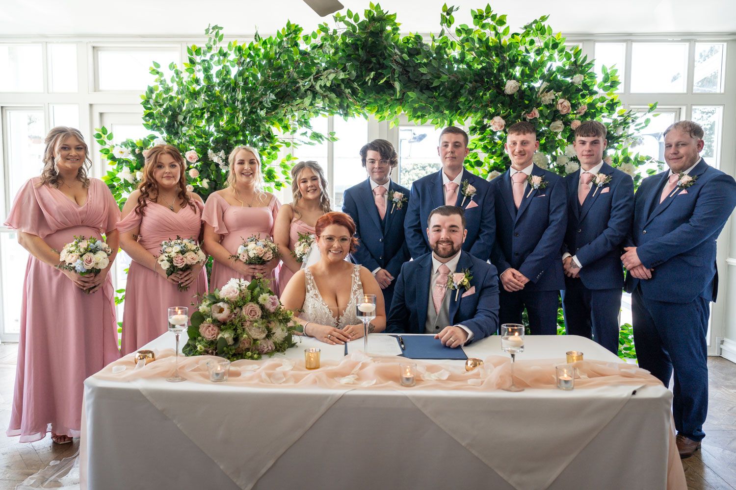 Wedding party gathers around couple at signing table with floral arch and pink bridesmaids dresses.