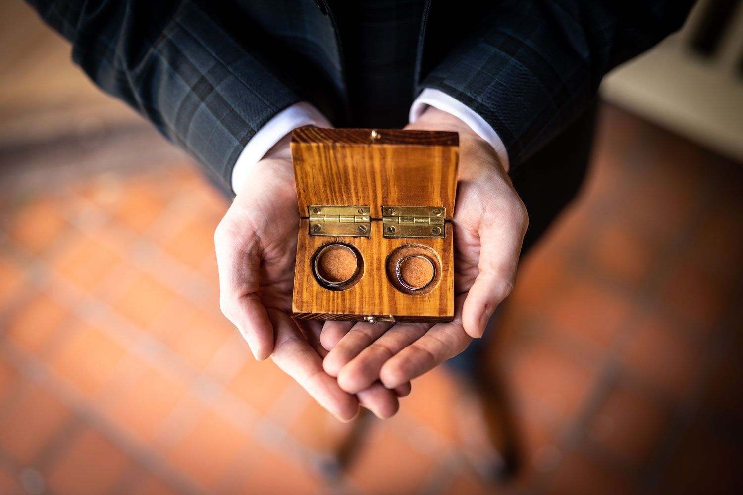 Hands holding small wooden box with two gold wedding rings displayed on red rustic tile floor.