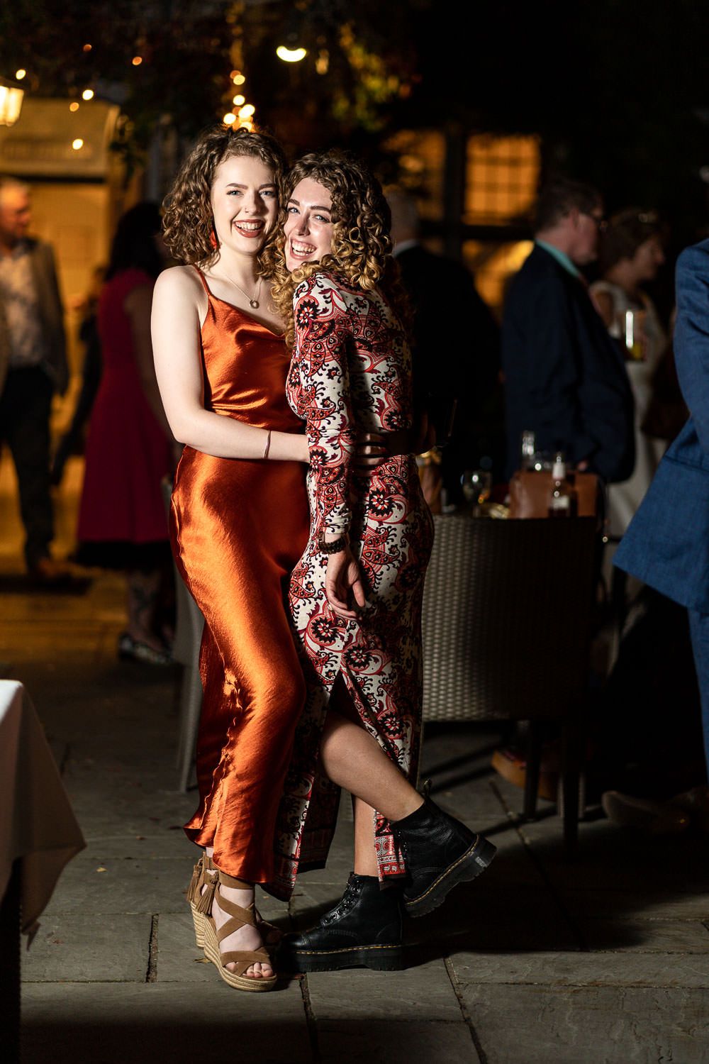 Two figures dancing together at a nighttime celebration, one wearing a rust-colored silk dress and one in a floral print.