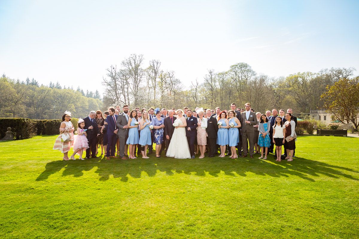 Large wedding party group photo on a sunny day standing together on bright green lawn with trees in background.
