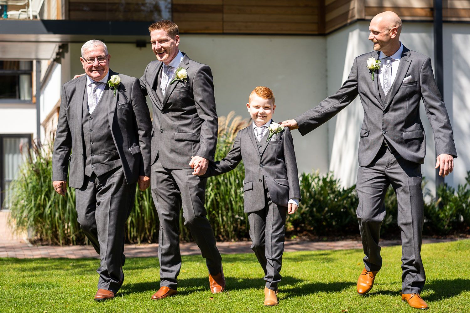 Four wedding party members wearing matching gray suits walk together on grass outside a building during a sunny day.