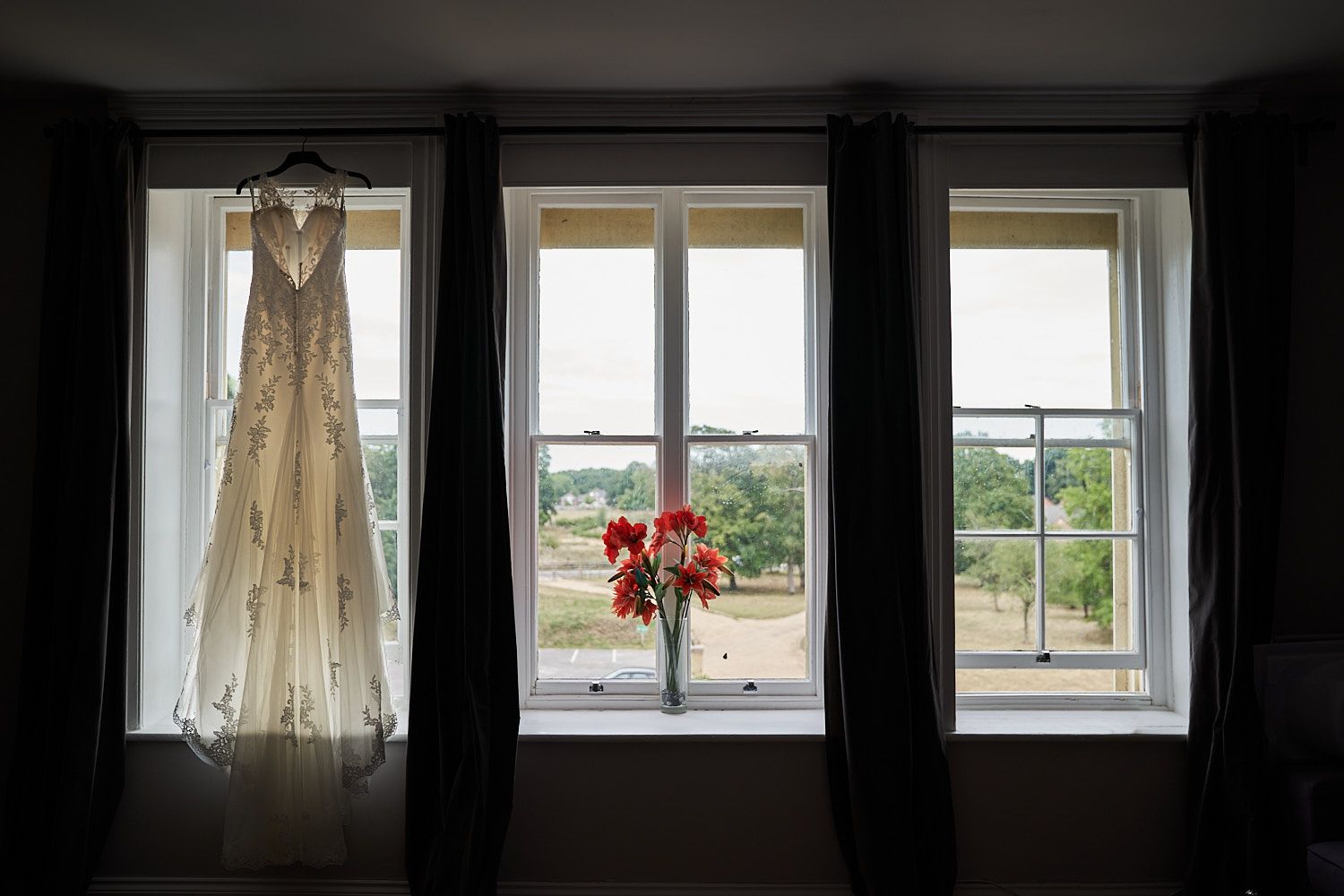 A white wedding dress hangs by a window with three panes overlooking green scenery and red flowers in a vase.