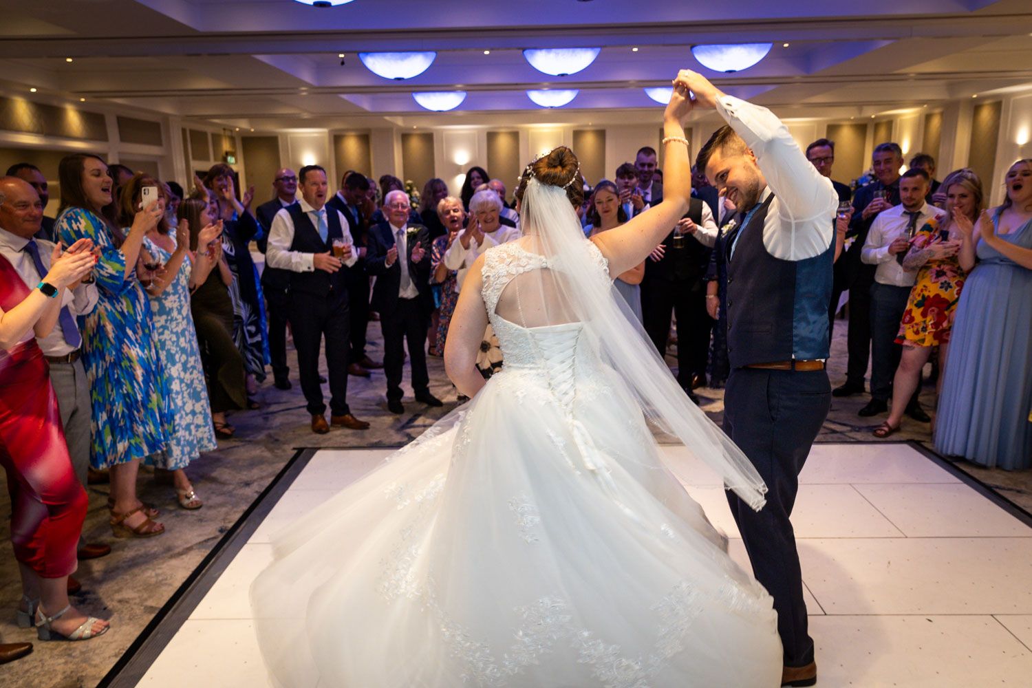 Wedding couple dancing surrounded by guests on illuminated reception dance floor.