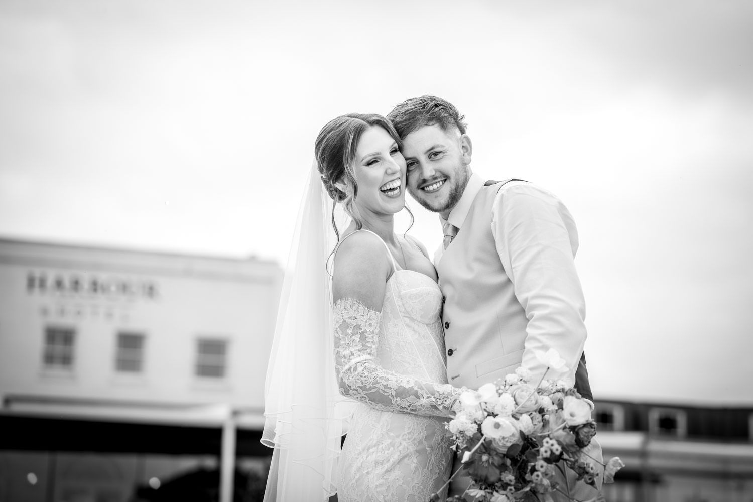 Happy bride and groom laugh together in black and white photo outside Harbour building.