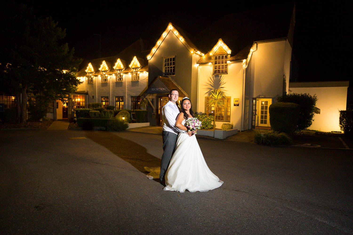 A couple in formal wedding attire embrace outside an illuminated venue building at night.