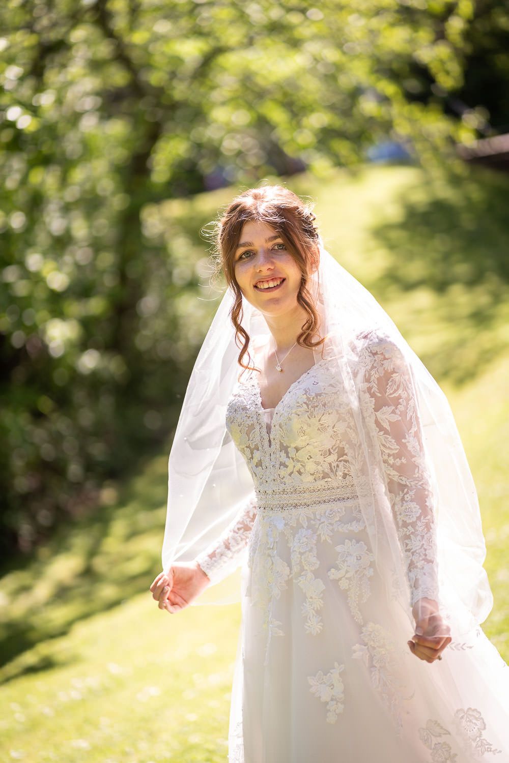 A bride wearing a long-sleeved lace wedding gown and veil stands in a sunny garden setting with bokeh background.