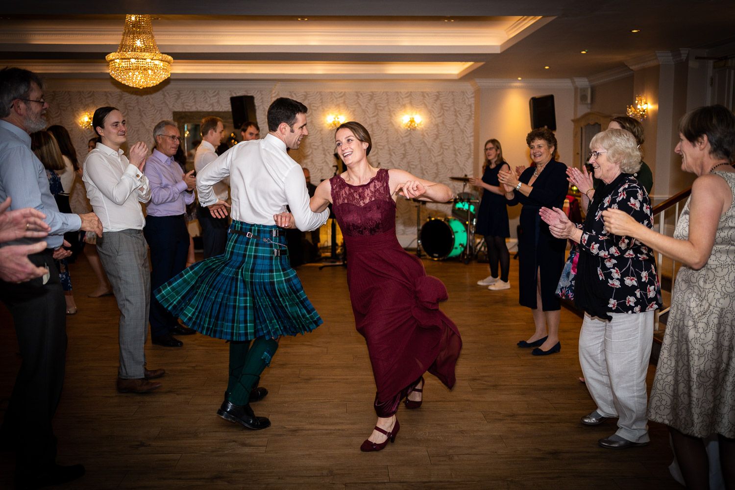 Wedding guests dance in a ballroom with a person wearing a traditional Scottish kilt among the dancers.