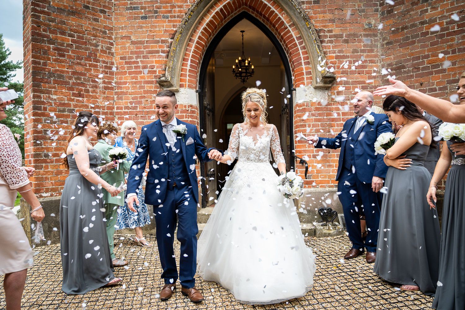 A newlywed couple exits a brick church as guests throw blue and white confetti during their wedding celebration.